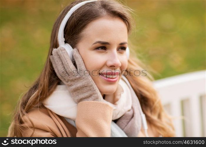 season, technology and people concept - beautiful happy young woman with headphones listening to music and sitting on bench in autumn park