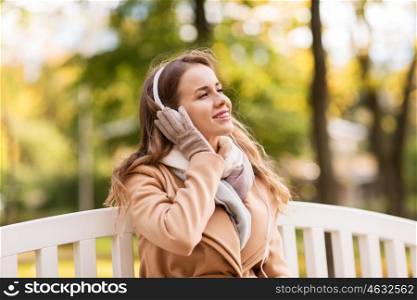 season, technology and people concept - beautiful happy young woman with headphones listening to music and sitting on bench in autumn park