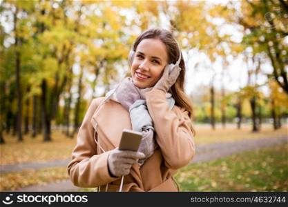 season, technology and people concept - beautiful happy young woman with headphones listening to music on smartphone walking in autumn park
