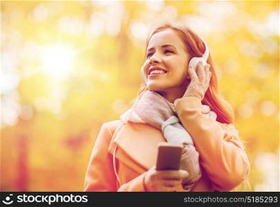 season, technology and people concept - beautiful happy young woman with headphones listening to music on smartphone walking in autumn park. woman with smartphone and earphones in autumn park