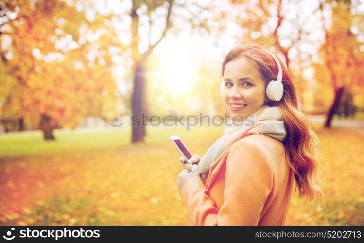 season, technology and people concept - beautiful happy young woman with headphones listening to music on smartphone walking in autumn park. woman with smartphone and earphones in autumn park