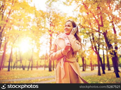 season, technology and people concept - beautiful happy young woman with headphones listening to music on smartphone walking in autumn park. woman with smartphone and earphones in autumn park