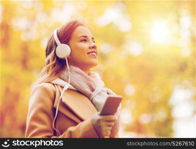 season, technology and people concept - beautiful happy young woman with headphones listening to music on smartphone walking in autumn park. woman with smartphone and earphones in autumn park
