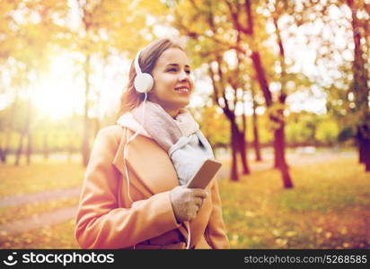 season, technology and people concept - beautiful happy young woman with headphones listening to music on smartphone walking in autumn park. woman with smartphone and earphones in autumn park