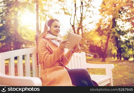 season, technology and people concept - beautiful happy young woman with tablet pc computer and headphones listening to music and sitting on bench in autumn park. woman with tablet pc and headphones in autumn park