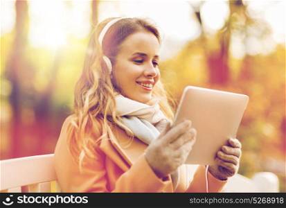 season, technology and people concept - beautiful happy young woman with tablet pc computer and headphones listening to music and sitting on bench in autumn park. woman with tablet pc and headphones in autumn park