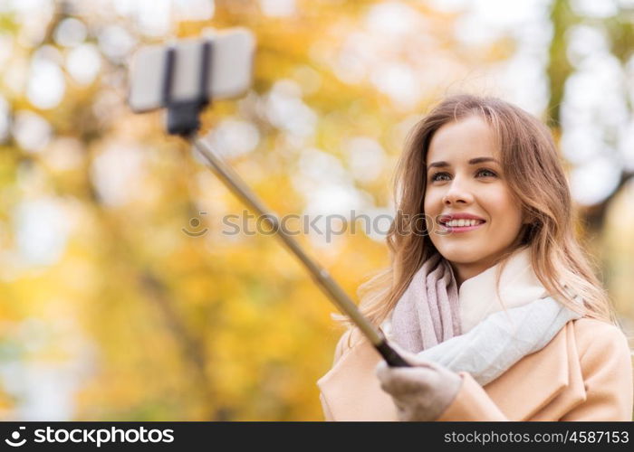 season, technology and people concept - beautiful young happy woman taking picture with smartphone selfie stick in autumn park