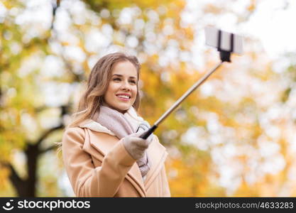 season, technology and people concept - beautiful young happy woman taking picture with smartphone selfie stick in autumn park