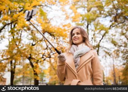 season, technology and people concept - beautiful young happy woman taking picture with smartphone selfie stick in autumn park