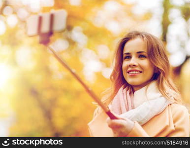 season, technology and people concept - beautiful young happy woman taking picture with smartphone selfie stick in autumn park. woman taking selfie by smartphone in autumn park