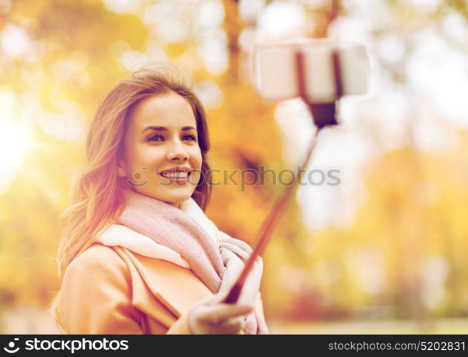 season, technology and people concept - beautiful young happy woman taking picture with smartphone selfie stick in autumn park. woman taking selfie by smartphone in autumn park