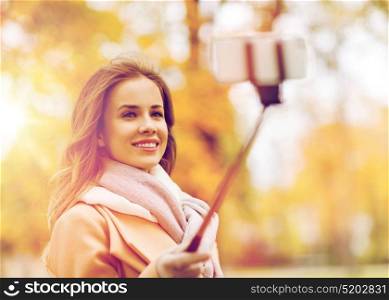 season, technology and people concept - beautiful young happy woman taking picture with smartphone selfie stick in autumn park. woman taking selfie by smartphone in autumn park