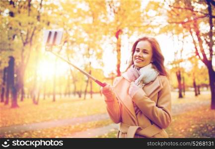 season, technology and people concept - beautiful young happy woman taking picture with smartphone selfie stick in autumn park. woman taking selfie by smartphone in autumn park