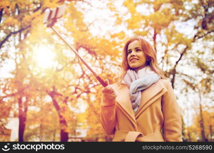 season, technology and people concept - beautiful young happy woman taking picture with smartphone selfie stick in autumn park. woman taking selfie by smartphone in autumn park