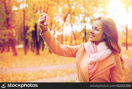 season, technology and people concept - beautiful young happy woman taking selfie with smartphone in autumn park. woman taking selfie by smartphone in autumn park