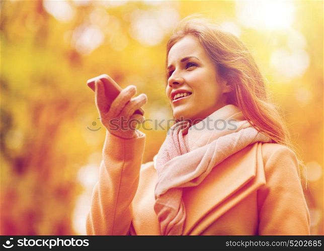 season, technology and people concept - beautiful young woman in autumn park and using voice command recorder on smartphone. woman recording voice on smartphone in autumn park