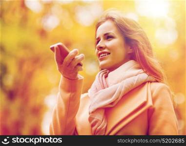 season, technology and people concept - beautiful young woman in autumn park and using voice command recorder on smartphone. woman recording voice on smartphone in autumn park