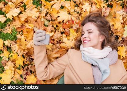 season, technology and people concept - beautiful young woman lying on ground and autumn leaves and taking selfie with smartphone