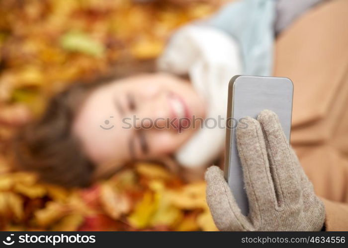 season, technology and people concept - beautiful young woman lying on ground and autumn leaves and taking selfie with smartphone