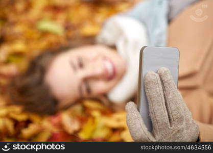 season, technology and people concept - beautiful young woman lying on ground and autumn leaves and taking selfie with smartphone