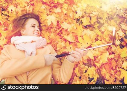 season, technology and people concept - beautiful young woman lying on ground and autumn leaves and taking picture with smartphone selfie stick. woman on autumn leaves taking selfie by smartphone