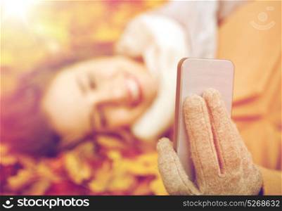 season, technology and people concept - beautiful young woman lying on ground and autumn leaves and taking selfie with smartphone. woman on autumn leaves taking selfie by smartphone