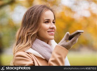 season, technology and people concept - beautiful young woman walking in autumn park and using voice command recorder on smartphone
