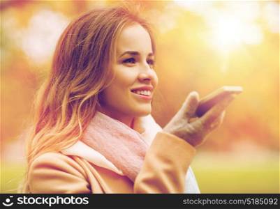 season, technology and people concept - beautiful young woman walking in autumn park and using voice command recorder on smartphone. woman recording voice on smartphone in autumn park