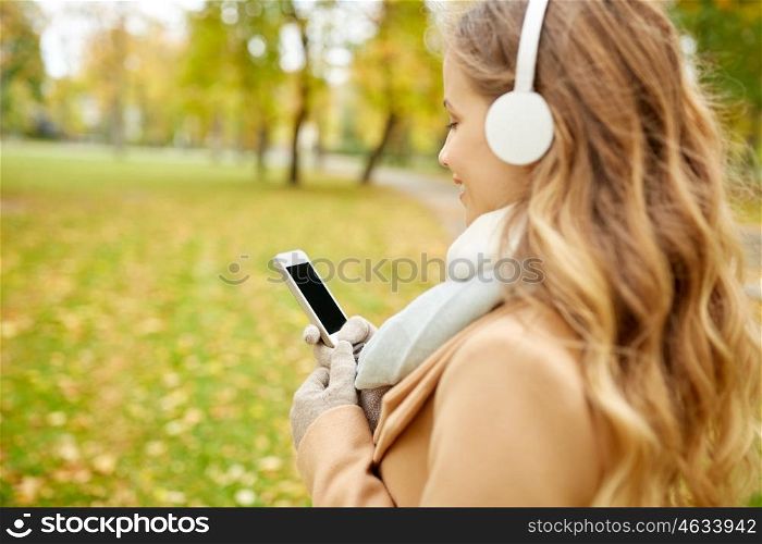 season, technology and people concept - close up of beautiful happy young woman with headphones listening to music on smartphone walking in autumn park