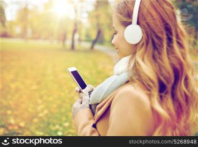season, technology and people concept - close up of beautiful happy young woman with headphones listening to music on smartphone walking in autumn park. woman with smartphone and earphones in autumn park