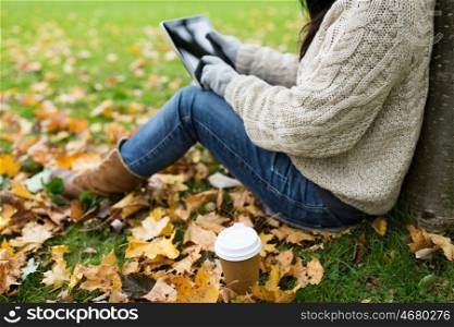 season, technology and people concept - young woman with tablet pc and coffee cup sitting on grass in autumn park