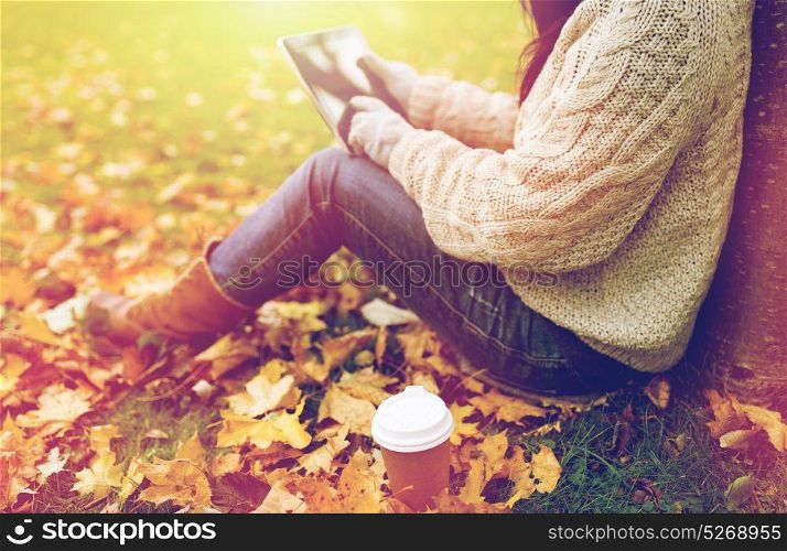 season, technology and people concept - young woman with tablet pc and coffee cup sitting on grass in autumn park. woman with tablet pc and coffee in autumn park