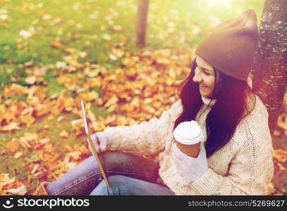 season, technology and people concept - young woman with tablet pc and coffee cup sitting on grass in autumn park. woman with tablet pc and coffee in autumn park