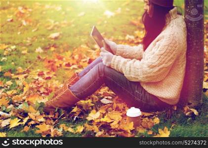 season, technology and people concept - young woman with tablet pc and coffee cup sitting on grass in autumn park. woman with tablet pc and coffee in autumn park