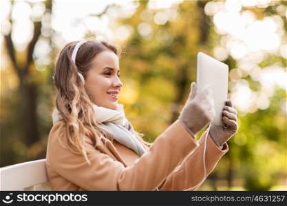season, technology, communication and people concept - beautiful happy young woman with tablet pc computer and headphones listening to music or having video call and sitting on bench in autumn park