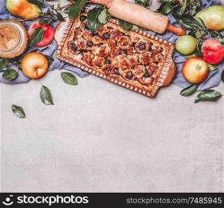 Seasonal garden fruits pie with rolling pin, ingredients, branches and leaves on gray background, top view. Border. Copy space for your design. Horizontal