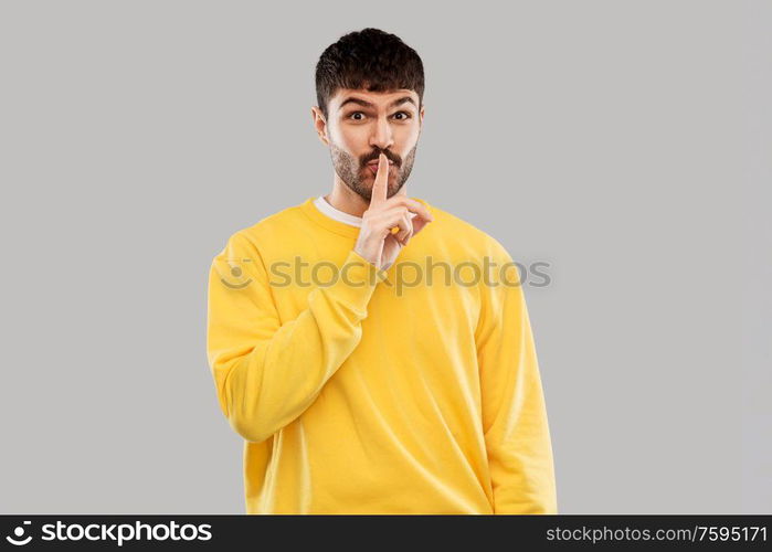 secret and people concept - smiling young man in yellow sweatshirt making hush gesture with finger on lips over grey background. young man making hush gesture with finger on lips