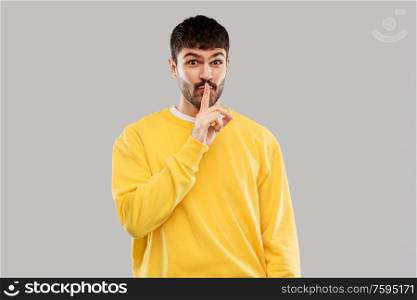 secret and people concept - smiling young man in yellow sweatshirt making hush gesture with finger on lips over grey background. young man making hush gesture with finger on lips