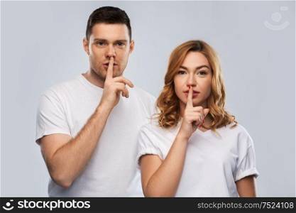 secret, conspiracy and people concept - portrait of happy couple in white t-shirts making hush gesture over grey background. happy couple in white t-shirts making hush gesture