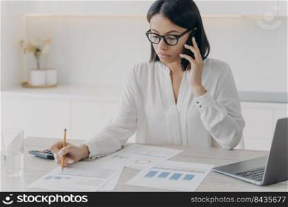 Secretary is appointing meeting on phone. Young european businesswoman in glasses and white blouse working remote from home. Manager or business assistant is sitting at the desk and doing research.. Secretary appointing meeting on phone. Business assistant is sitting at the desk and doing research.