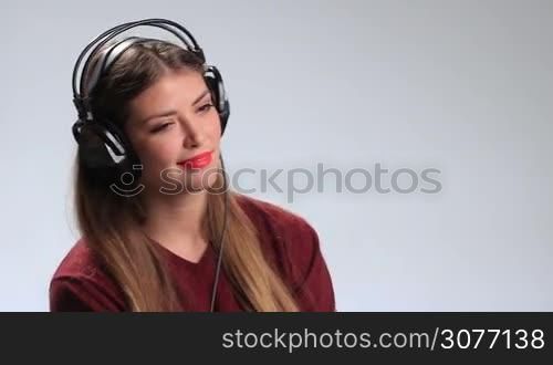 Seductive young brunette woman wearing red lipstick and listening to music in headphones. Sexy pretty girl with playful expression, twisting long hair around her finger and looking at the camera teasingly while enjoying favorite track in earphones.