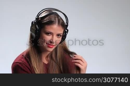 Seductive young brunette woman wearing red lipsticks and listening music in headphones. Playful sexy girl twisting her beautiful long hair around her finger, looking at camera temptingly and alluringly while enjoying her favorite track in earphones.