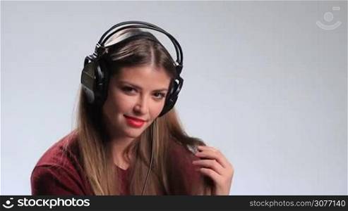 Seductive young brunette woman wearing red lipsticks and listening music in headphones. Playful sexy girl twisting her beautiful long hair around her finger, looking at camera temptingly and alluringly while enjoying her favorite track in earphones.