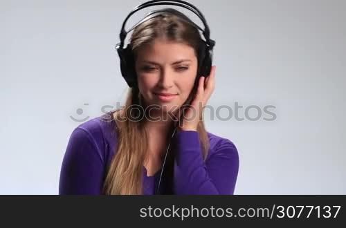 Seductive young brunette woman with long hair listening track on mp3 player on white. Happy smiling girl in earphones enjoying music, touching one earcup, hearing the song fading, slowly taking off headphones and hanging them around her neck.