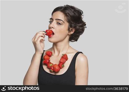 Seductive young woman wearing strawberry necklace as she eats one piece over gray background