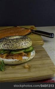 Seeded bagel with smoked salmon, cream cheese, cucumber, dill and capers on wooden board, half side in background, portrait copyspace to top.