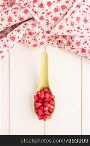 Seeds of red ripe peeled pomegranate on ceramic spoon. Rustic wood board background with fabric with little flowers dot fabric. Top view, copy space