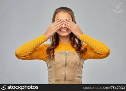 seeing, vision and people concept - smiling teenage girl closing her eyes by hands over grey background. smiling teenage girl closing her eyes by hands