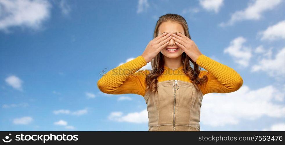 seeing, vision and people concept - smiling teenage girl closing her eyes by hands over blue sky and clouds background. smiling teenage girl closing her eyes over sky