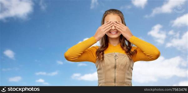 seeing, vision and people concept - smiling teenage girl closing her eyes by hands over blue sky and clouds background. smiling teenage girl closing her eyes over sky